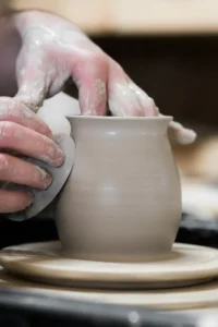 Een pot in witte klei op de pottenbakkers schijf, handen houden een metalen lomer vast